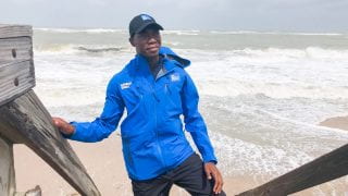 Tevin Wooten speaking on air of The Weather Channel during a storm with a beach line and ocean in the background.